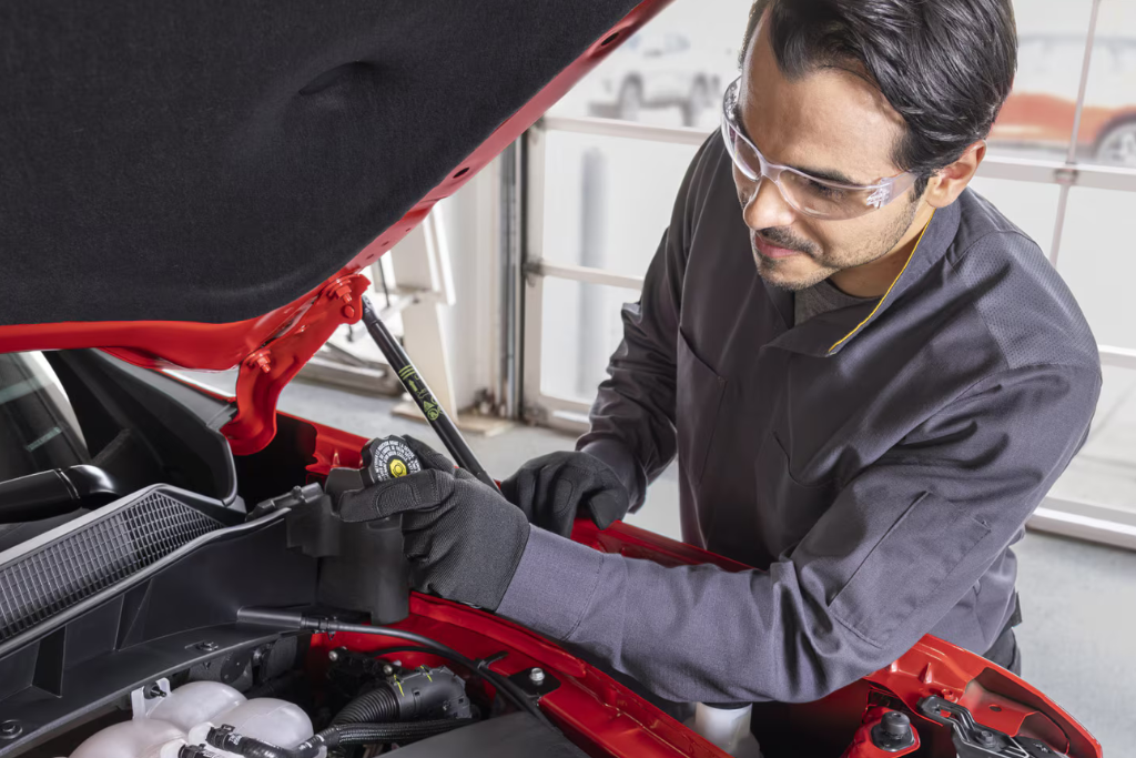 Mechanic inspecting coolant reservoir under the hood of a red Chevrolet vehicle, wearing safety goggles and gloves in a service bay.
